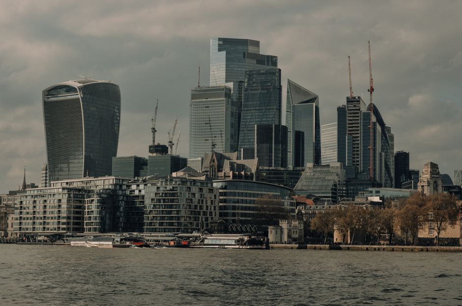 View of a modern city skyline along the River Thames, featuring tall glass skyscrapers including one with a curved design on the left and another with a stepped, angular appearance on the right. In the foreground, there are mid-rise residential buildings with balconies, situated adjacent to the riverbank. Several boats and watercraft are seen navigating the water near the shoreline. Construction cranes are visible atop some of the skyscrapers, indicating ongoing development. The scene is captured during daylight with overcast skies, providing natural lighting that highlights the reflective surfaces of the high-rise buildings. This urban landscape suggests a central business district area, relevant to home relocation and furniture transport activities associated with moving services, exemplified by the proximity to residential and commercial properties near the water. The image aligns with themes of city-based relocations and logistical planning for house removals serviced by Removal Companies Waterloo.