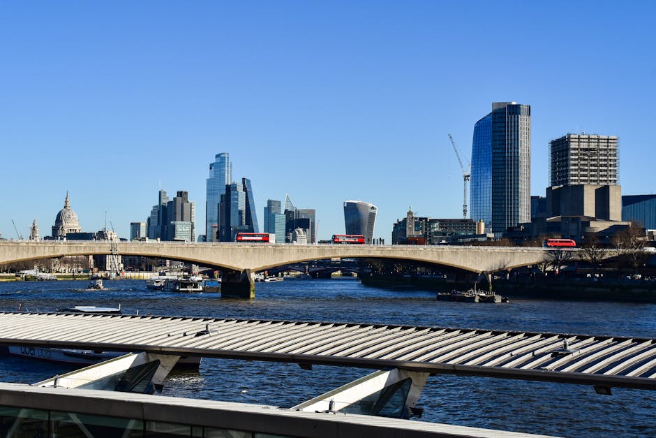 View of a modern city skyline along the River Thames, featuring tall glass skyscrapers including one with a curved design on the left and another with a stepped, angular appearance on the right. In the foreground, there are mid-rise residential buildings with balconies, situated adjacent to the riverbank. Several boats and watercraft are seen navigating the water near the shoreline. Construction cranes are visible atop some of the skyscrapers, indicating ongoing development. The scene is captured during daylight with overcast skies, providing natural lighting that highlights the reflective surfaces of the high-rise buildings. This urban landscape suggests a central business district area, relevant to home relocation and furniture transport activities associated with moving services, exemplified by the proximity to residential and commercial properties near the water. The image aligns with themes of city-based relocations and logistical planning for house removals serviced by Removal Companies Waterloo.